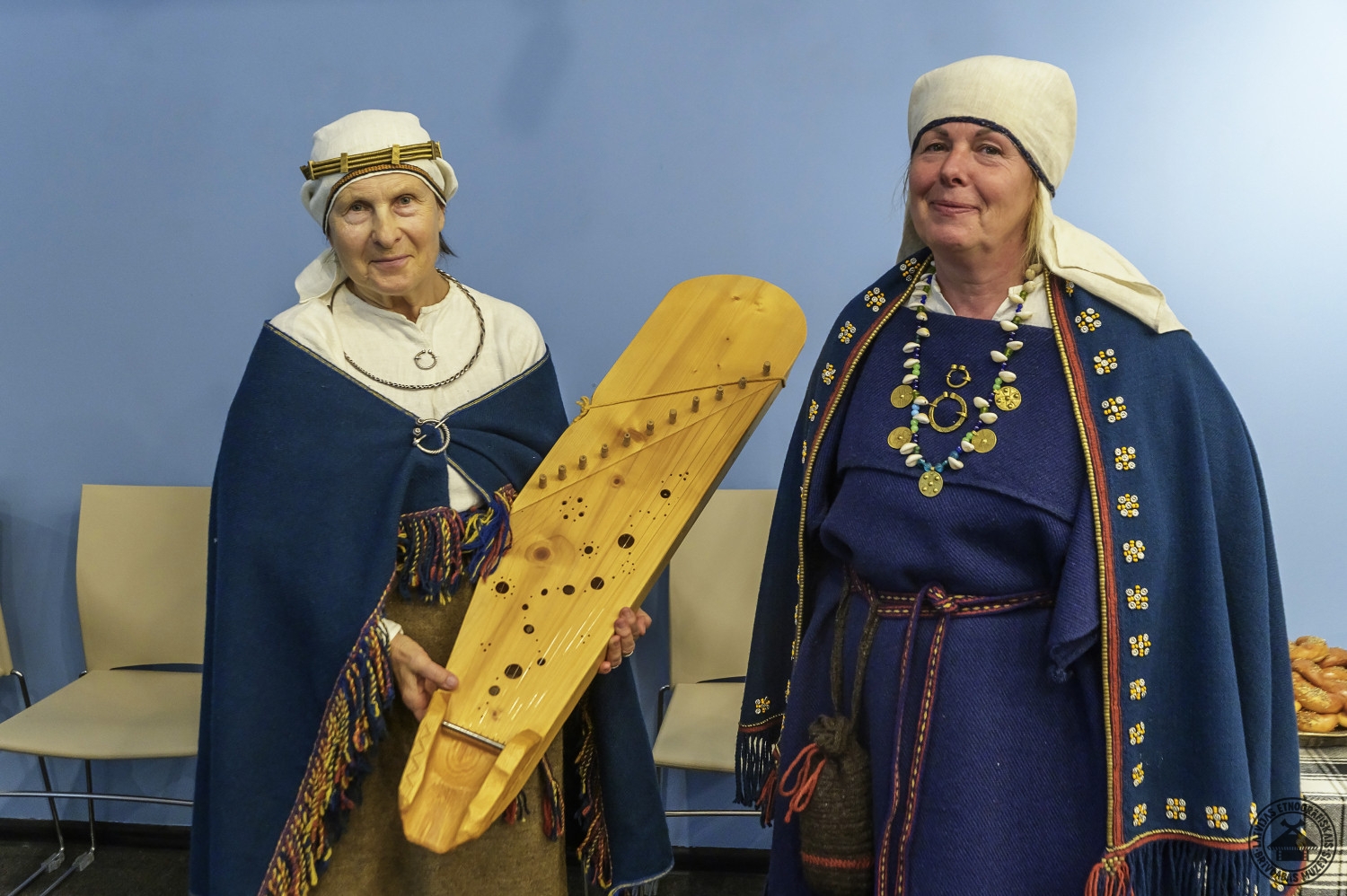 Divas sievietes tautastērpos; viena no viņām tur rokās latviešu etnogrāfisko kokli. 