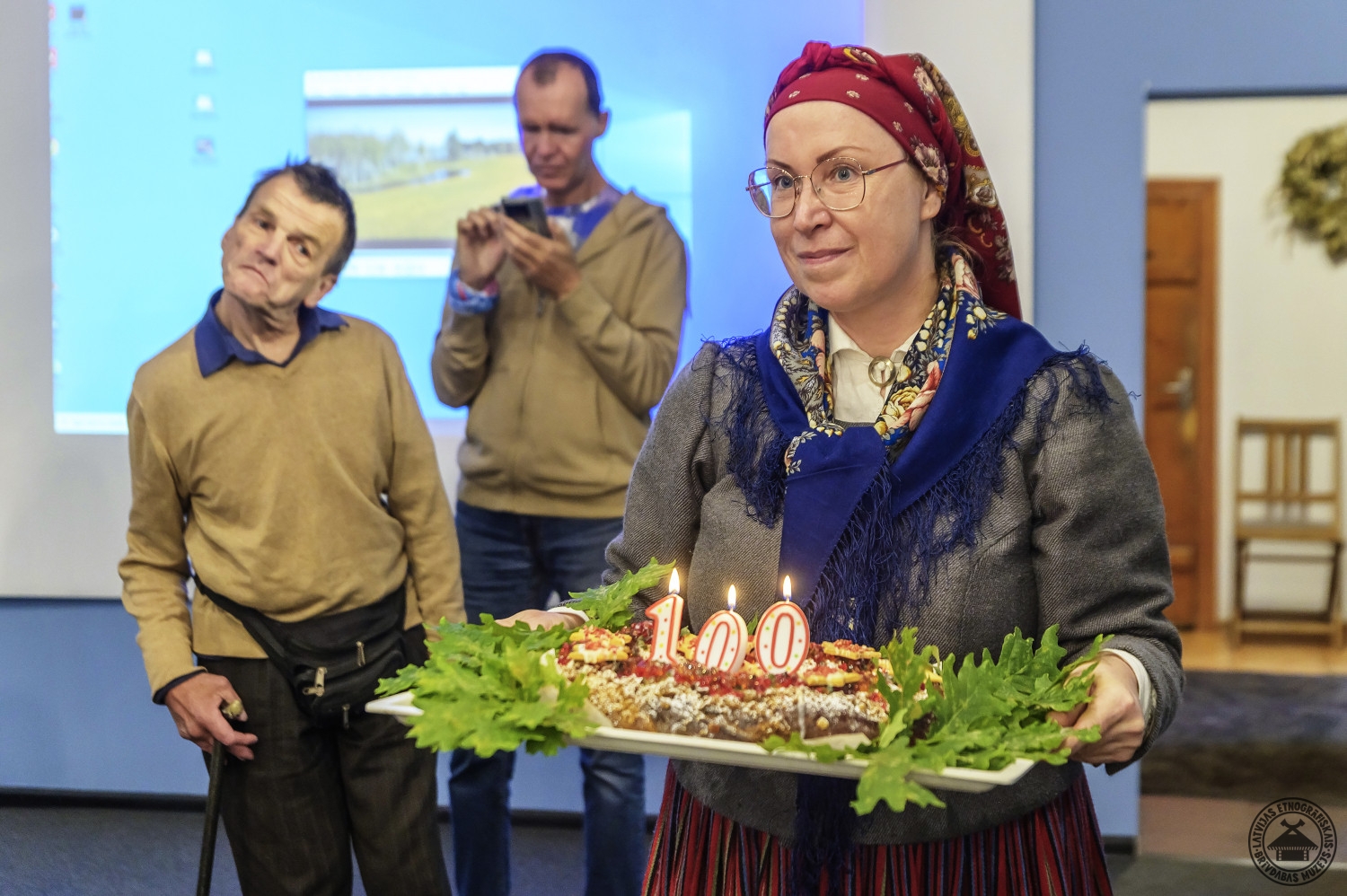 Muzeja darbiniece tautastērpā nes svētku kliņģeri ar jubilejas svecēm “100”  