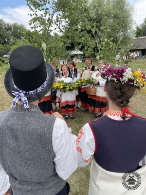 Jaunais pāris stāv pretī Rucavas sievām, kuras rokās tur pītu ziedu un zaļumu virteni. 