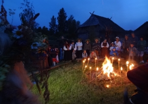 Jāņu ugunskurs "Vītolnieku" sētā, ap kuru iedegtas lāpas un stāv svinētāji.