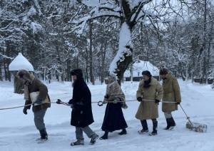 Cilvēki sniegainā lauku ainavā, tērpušies siltās seno laiku drēbēs, ar spēku velk lielu koka bluķi