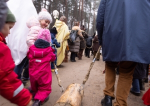 Bluķa vilkšanas gājiens, kurā tuvplānā redzams liels, raupjš koka bluķis, kas tiek vilkts pa sniegu