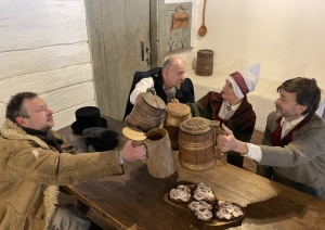 Trīs kungi un sieviete tautastērpos sēž pie koka galda ar alus kausiem, svinot Teņa dienu