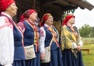 Četras sievas Rucavas tautastērpos stāv pie vēsturiskas klētiņas un dzied tautasdziesmas.