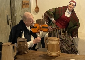 Muzikāls priekšnesums: vīrietis spēlē vijoli, kamēr sieviete tautastērpā viņa pavadījumā dejo