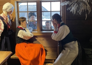 Meitas tautastērpos pie loga nepacietīgi gaida ciemos nākam puišus; fonā sniegota ziemas ainava