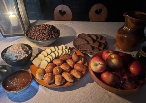 Tradicionālais veļu mielasts māla un koka traukos uz koka galda: tuvplānā redzami pīrāgi, biezputra, pelēkie zirņi, āboli, rupjmaize un siers Latvijas Etnogrāfiskā brīvdabas muzeja klētī.