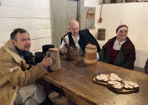 Svētku viesi krogā sēž pie gariem koka galdiem, malkojot alu no tradicionāliem kausiemSvētku viesi krogā sēž pie gariem koka galdiem, malkojot alu no tradicionāliem kausiem