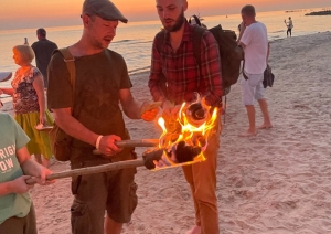 Cilvēki ar aizdegtām lāpām stāv jūras krastā uz krāšņa vakara saulrieta fona.